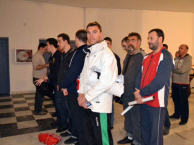 Un grupo de hombres en una sala con una pared de azulejos y una puerta azul. Algunos hombres están de pie y otros están sentados.