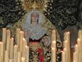 Una estatua religiosa con un vestido de estilo barroco y un tocado dorado. Alrededor de la estatua hay varios velas encendidas, iluminando el ambiente. La escena parece ser parte de una procesión o celebración religiosa, con un fondo decorado en tonos oscuros.