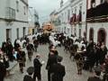 Un desfile militar en una calle adoquinada con edificios blancos y rojos. Soldados vestidos de uniforme negro, sosteniendo armas y banderas, avanzan por el camino. La atmósfera es formal y ceremonial.