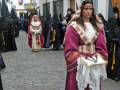 Imagen muestra a una mujer vestida con traje tradicional, posiblemente durante una procesión o celebración cultural. El entorno parece ser urbano con edificios blancos y calles adoquinadas.