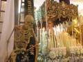 Una escultura de esqueleto humano se encuentra en el centro del imagen, rodeado por velas encendidas y flores. La escultura parece estar en una iglesia o lugar de culto, con detalles arquitectónicos y decoraciones visibles en el fondo.