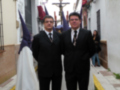 Dos hombres en trajes posan frente a un crucifijo, con una figura vestida de blanco y capucha en el fondo. La escena se desarrolla en un entorno urbano con edificios y calles típicas.
