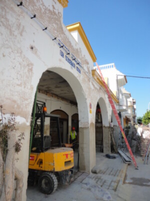 En construcción, una grúa amarilla trabaja en la renovación de un edificio histórico con arcos y azulejos. La estructura muestra signos de deterioro, mientras que el cielo azul despejado brilla sobre la obra.