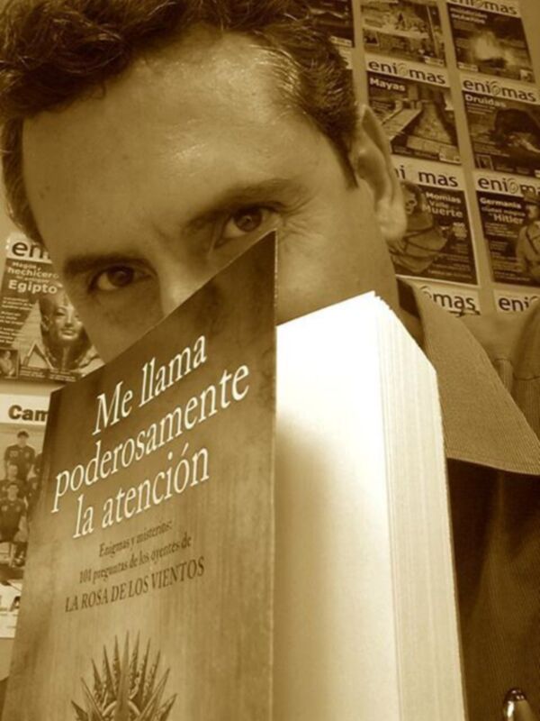 Un hombre con el rostro oculto detrás de un libro abierto que muestra la portada "La Rosa de los Vientos" con el título "Me llama poderosamente la atención". El libro está en una pared de periódicos.