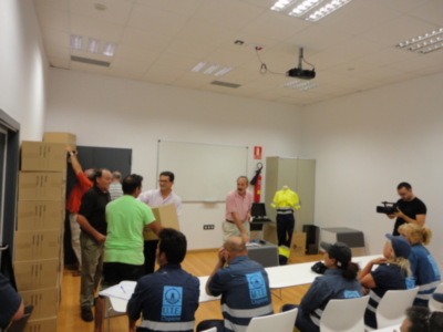 En un aula iluminada con cajas de cartón, personas interactúan en torno a una mesa. Algunos llevan uniformes de trabajo, mientras otros observan la actividad. La imagen sugiere un entorno de aprendizaje o capacitación en un espacio educativo.