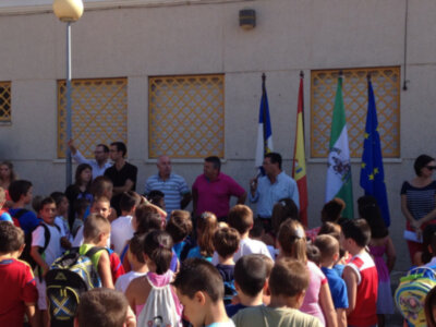 Un grupo de niños y adultos están reunidos frente a una pared con ventanales amarillos. Algunos niños llevan mochilas y camisetas de colores, mientras otros adultos observan la reunión. En el fondo, se pueden ver banderas de diferentes países, incluyendo la española y la europea. La escena parece ser una actividad escolar o de comunidad en un entorno urbano.