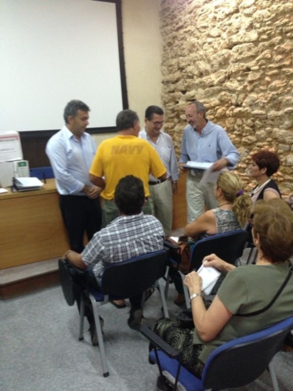 En una sala de conferencias, un grupo de personas se reúne alrededor de una mesa con documentos. La pared de fondo es de piedra, y en el extremo izquierdo se ve un escritorio con una computadora portátil. La atmósfera parece ser de trabajo o reunión formal.