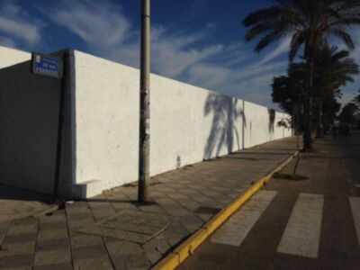 Pared blanca con señal de calle 'Avenida 28 de Febrero' junto a una acera adoquinada con palmeras al fondo bajo un cielo azul con nubes.