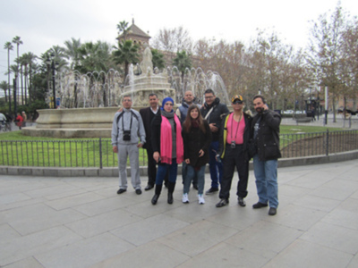En un parque con palmeras y una fuente, seis personas posan frente a la cámara. Algunos llevan cascos y otros son fotógrafos, capturando momentos en la ciudad.