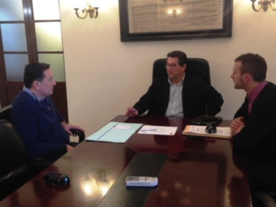 Tres hombres en una reunión de negocios, sentados frente a un escritorio con documentos y teléfonos móviles. La pared detrás de ellos tiene un marco con texto y dos lámparas colgantes.