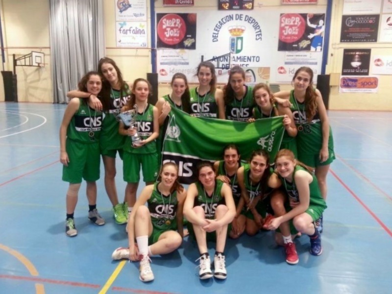 Equipo femenino de baloncesto posando con trofeo y bandera en cancha de baloncesto.