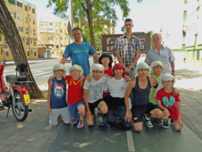 Un grupo de niños y adultos posan en una calle con árboles y edificios al fondo. Algunos llevan gorras blancas, otros camisetas y pantalones cortos. En la esquina hay una motocicleta roja.