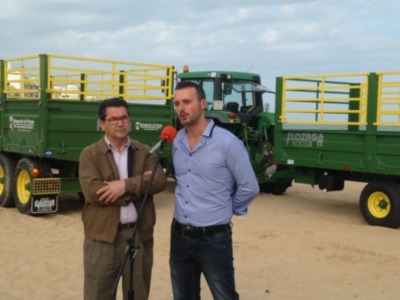 Dos hombres charlan en un campo de tierra, con un tractor y una remolque detrás.
