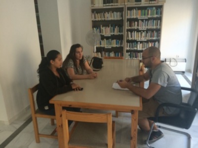 Tres personas sentadas en una mesa en una biblioteca, con libros y una ventiladora en el fondo.