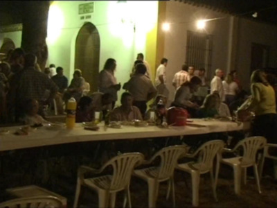 Una noche de fiesta en una terraza con gente reunida alrededor de mesas y sillas blancas. La iluminación es natural, proveniente de luces colgantes y una luz en la pared. La atmósfera es festiva, con muchos participantes disfrutando de la noche.