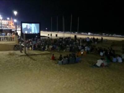 Una multitud disfruta de una película al aire libre en la playa, con un gran proyector y pantalla en el centro. La noche está oscura, iluminada por farolas y luces de la ciudad en el fondo. La arena está llena de espectadores sentados o acostados, todos disfrutando del evento.