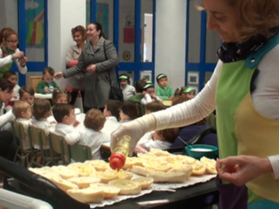 En una escuela infantil, niños y adultos están sentados en mesas. Una persona prepara pasteles en un estante de cocina mientras otros niños observan atentamente. La escena transmite una atmósfera de aprendizaje y colaboración en un entorno educativo.