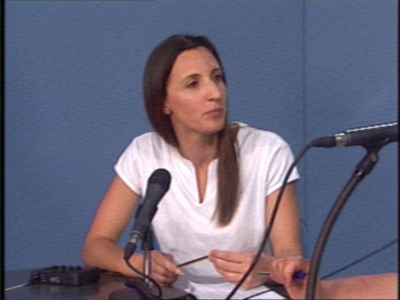 Persona hablando en un evento con micrófono y material de presentación en la mesa.