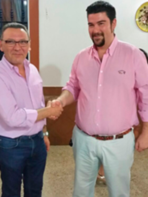 Tres hombres en una reunión formal, dos de ellos saludándose con una mano. El ambiente parece ser un lugar de trabajo o reunión, con elementos decorativos y una mesa en el fondo.