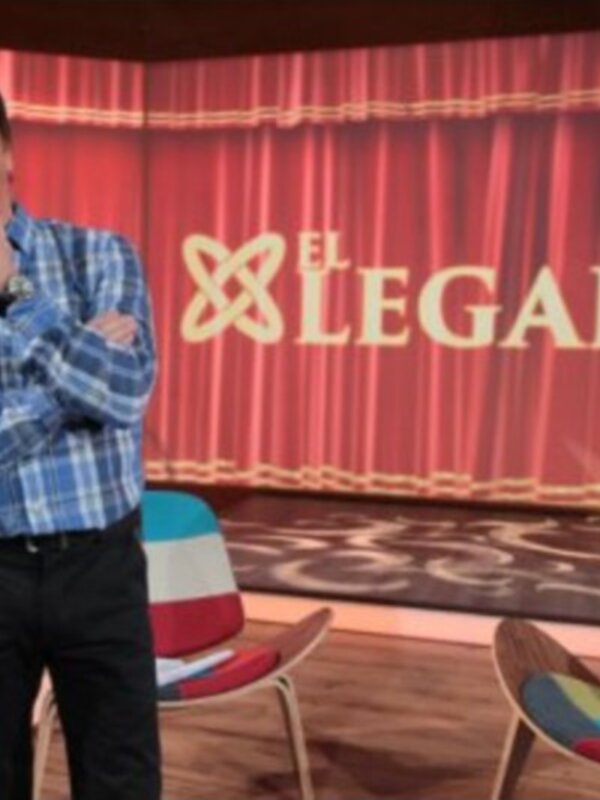 Un hombre con una camisa azul y pantalones negros está en un escenario de televisión, con una silla colorida al lado. El fondo es rojo con el texto "El Legado de..." en letras grandes y un logotipo.