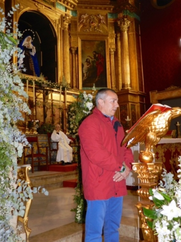 En una iglesia con decoración dorada y roja, un hombre en chaqueta roja y pantalones azules mira hacia la cámara. Detrás de él, una imagen religiosa con flores blancas y un altar dorado.