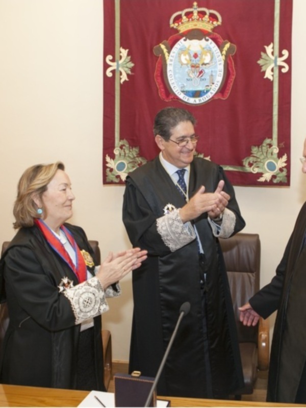 Un evento formal con tres personas en trajes formales, dos de ellas aplaudiendo a la tercera, frente a una bandera con un escudo heráldico. La escena sugiere un momento de reconocimiento o celebración.