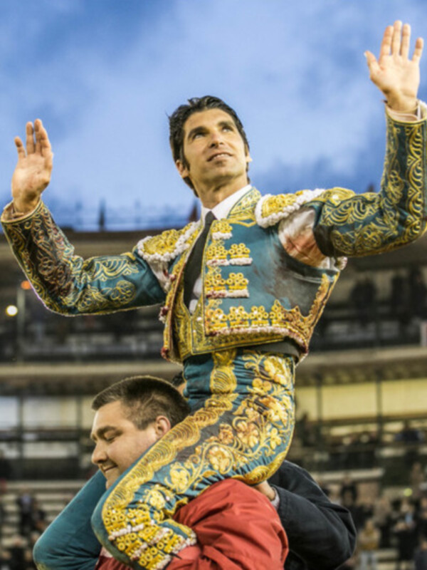 Un torero en traje tradicional sostiene a un hombre que parece estar en su espalda, durante una corrida de toros. La imagen se toma en una arena con asientos llenos y luces brillantes iluminando el escenario.