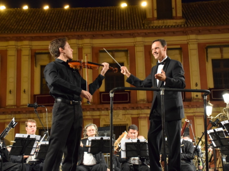 Concierto de orquesta en un teatro histórico. Dos violinistas se destacan, uno tocando y el otro dirigiendo con un bastón. La orquesta está formada por músicos en trajes de gala, todos concentrados en su interpretación. La atmósfera es intensa y el escenario iluminado destaca la seriedad del evento.