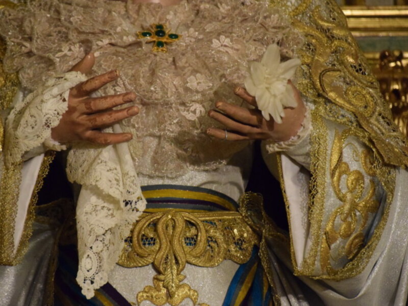 Una figura vestida con un atuendo ceremonial elaborado, probablemente de una celebración religiosa o festiva. El vestido está adornado con detalles dorados y oro, incluyendo una corona en la cabeza. La figura sostiene un objeto blanco en su mano izquierda, que parece ser una flor o un símbolo importante. La escena se enmarca en un entorno que sugiere una iglesia o templo, con elementos de dorado y ornamentación visible en el fondo.
