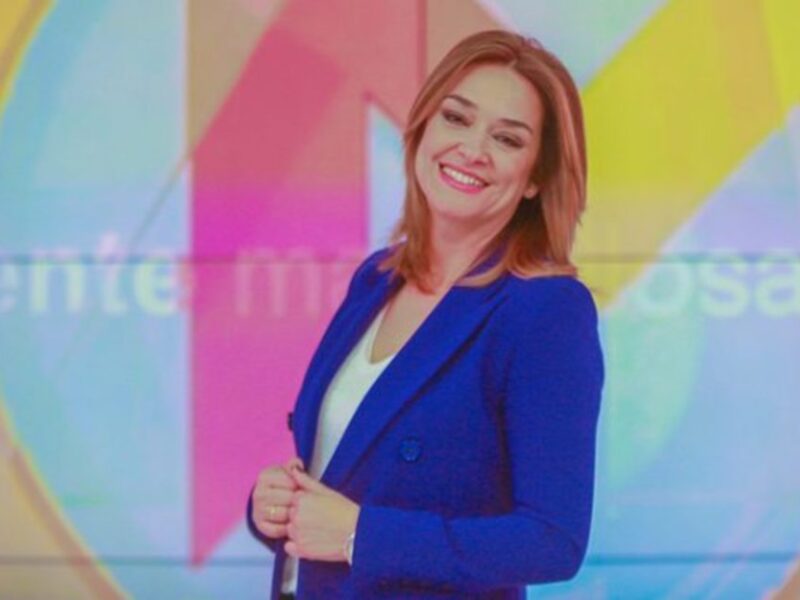 A una mujer con cabello largo y ondulado, sonriendo mientras sostiene un micrófono. El fondo es de colores brillantes y abstractos, con letras en la parte inferior que parecen ser "Gente" y "muy". La mujer está vestida con un blazer azul y una camiseta blanca.