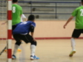 Jugadores en acción durante un partido de fútbol sala, con una portería visible y el suelo del gimnasio.