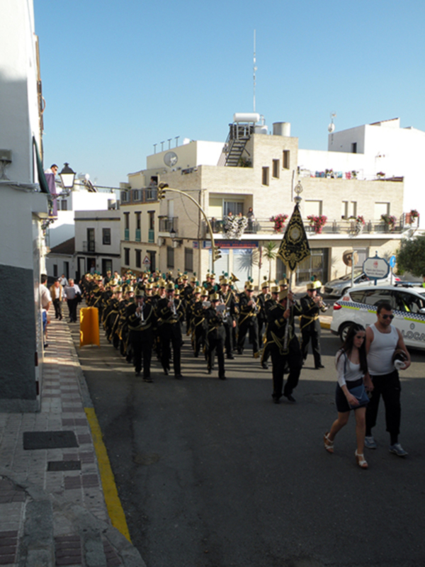 Desfile militar en una calle con edificios blancos y azules en el fondo, acompañado por una policía en el fondo.