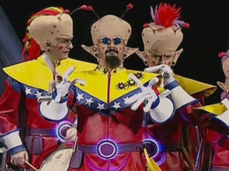 Actores en trajes coloridos con detalles estelares y máscaras antropomórficas en un escenario teatral.