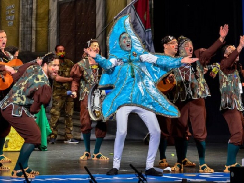 Un actor con una capa azul brillante y pantalones blancos está en el centro de la imagen, con un gesto dramático. A su alrededor, otros actores vestidos de colores diferentes están en pie o en movimiento. La escena parece ser parte de una actuación teatral, con un fondo que sugiere un escenario interior.