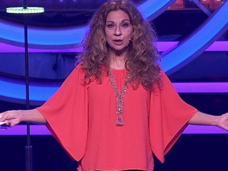 Una mujer con cabello largo y ondulado, usando una blusa roja y una gargantilla, se encuentra en una escena de televisión con un fondo azul y blanco.