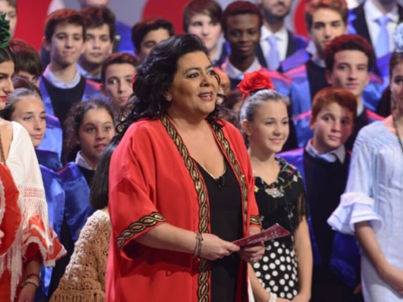 Una mujer en un vestido rojo con una corona de flores, frente a una audiencia de jóvenes vestidos formalmente.