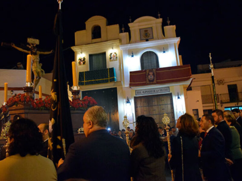 Imagen nocturna de una procesión religiosa en un pueblo español. En primer plano, una estatua de Jesús crucificado y una bandera negra. Al fondo, un edificio con detalles arquitectónicos y luces que iluminan la escena. La gente observa atentamente el evento, algunos con sombrillas para protegerse del viento.