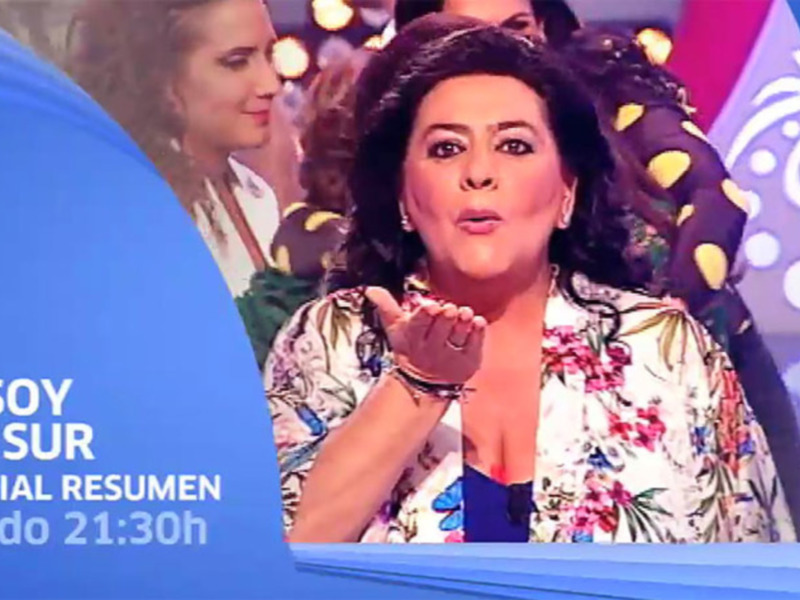 Una mujer con vestido floral y tocado de flores, en un escenario de televisión con fondo azul y amarillo.