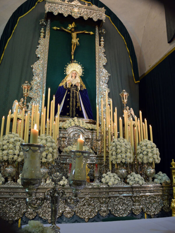 Un altar religioso con una imagen de la Virgen María y el Cristo crucificado, rodeado por flores blancas y velas encendidas. La decoración es elaborada con detalles dorados y cristales, en un ambiente de armonía y reverencia.