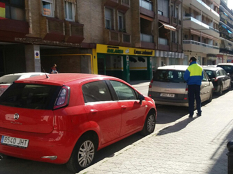 Fotografía de un estacionamiento en una calle de la ciudad. En primer plano, un coche rojo y en segundo plano, varios vehículos estacionados. A la derecha, un policía en uniforme de trabajo observa el estacionamiento. En el fondo, edificios con ventanas y un letrero amarillo de una tienda. La imagen muestra un día soleado en la ciudad, con sombras caídas de los edificios.