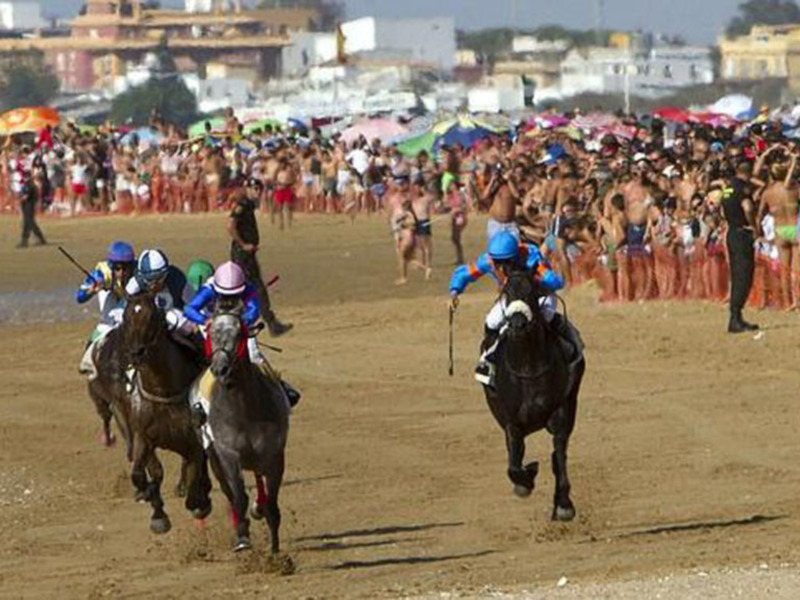 Corrida de caballos en la playa, con jinetes en acción y espectadores al borde del agua.