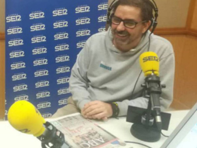 Un hombre sonríe mientras está en una radio, con un micrófono y un periódico en la mesa. La pared detrás de él tiene el logo de "SER 12".