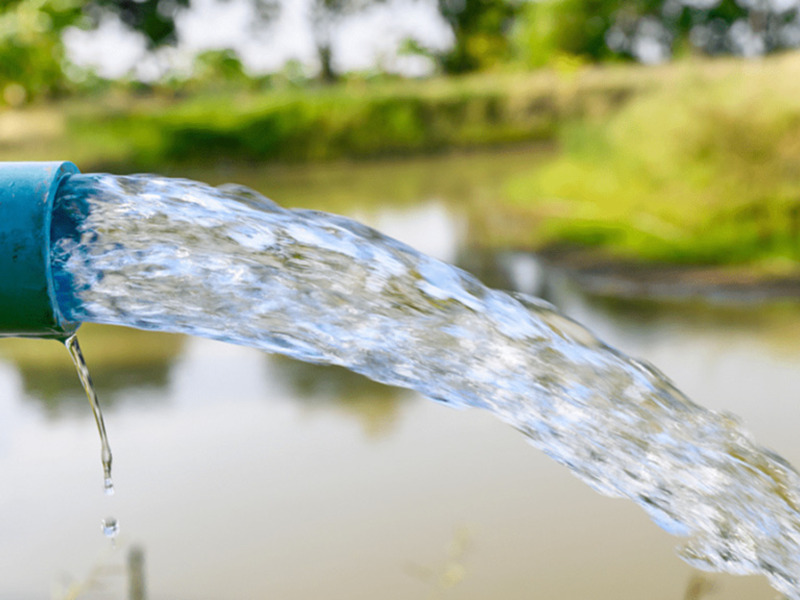 Agua fluindo de un tubo hacia un estanque, con vegetación y cielo despejado en el fondo.