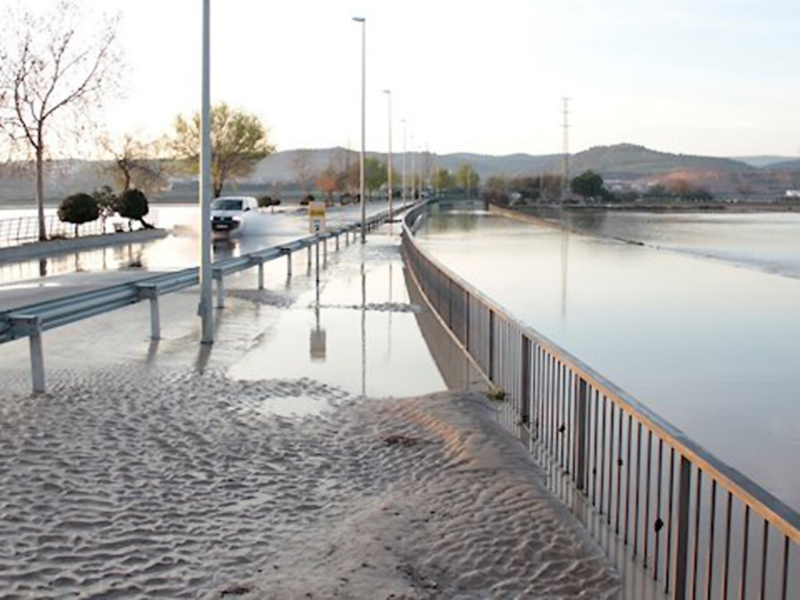 Un área de agua tranquila rodeada por una valla metálica y arena. Hay algunos árboles secos en el fondo, con montañas lejanas y una línea de horizonte clara. La escena sugiere un lugar tranquilo para caminar o relajarse cerca del agua.