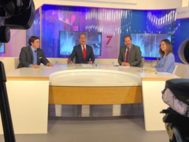 Un estudio de televisión con tres personas sentadas en una mesa, frente a un fondo con pantallas y el número 7. Una cámara de televisión está enfocando la escena.