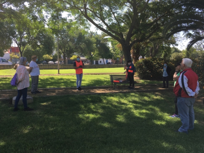 Una imagen de un grupo de personas en un parque, algunos con chalecos reflectantes. En el fondo hay árboles y edificios, con un cielo azul claro. La gente parece estar en una actividad o reunión al aire libre.
