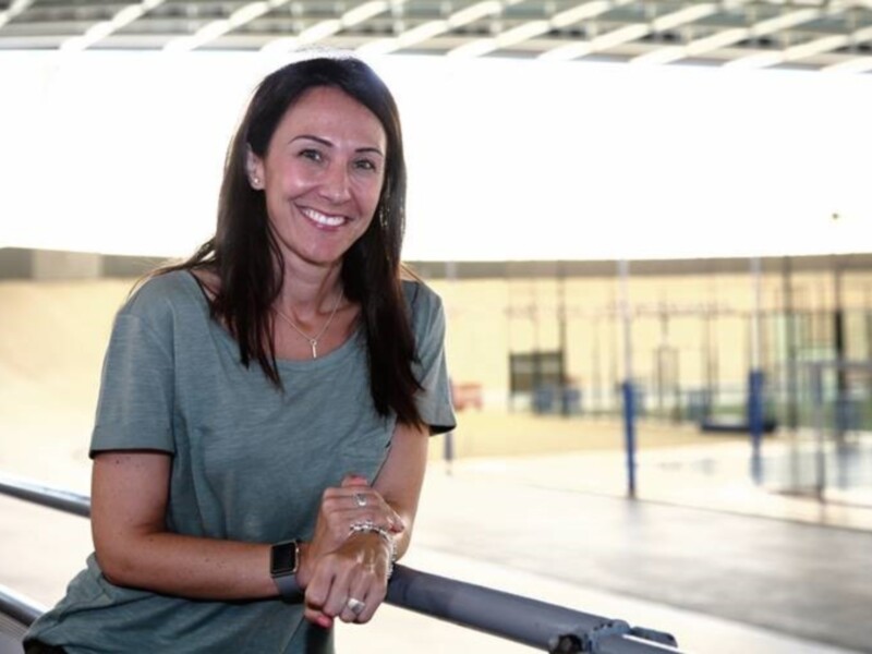 A una mujer sonriendo mientras sostiene un objeto en la mano. El fondo es una estructura moderna con cristales y metal, probablemente un edificio o centro comercial.