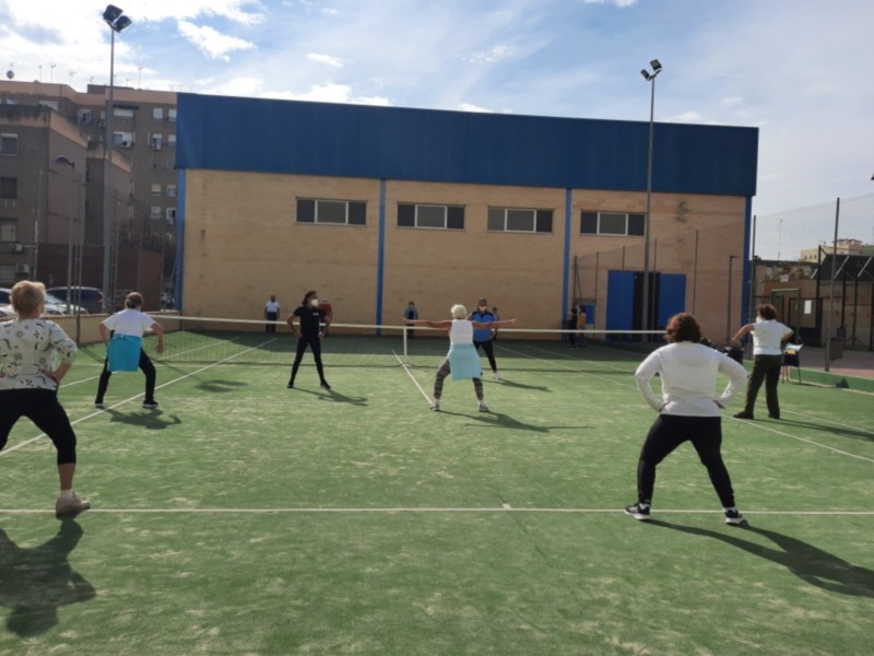 Un grupo de personas en una cancha de tenis, con un edificio de fondo y dos postes de luz.
