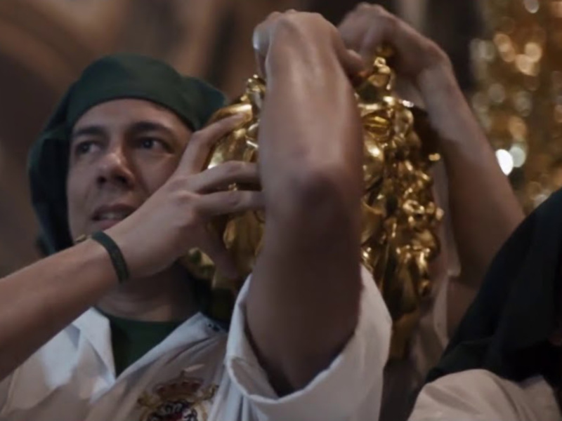 Imagen de un hombre con una gorra verde sujetando una corona dorada, mientras otros participantes observan. Contexto: ceremonia o celebración religiosa.