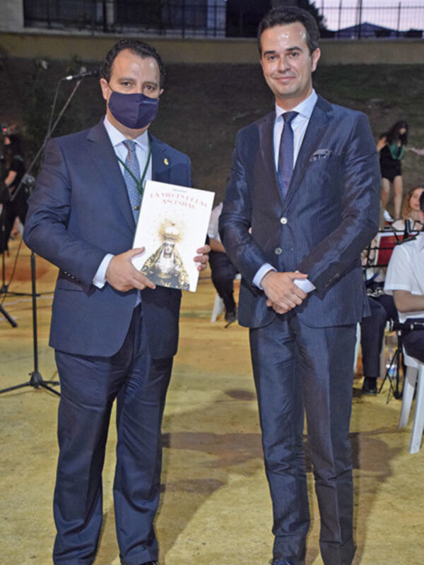 Dos hombres en trajes azules posan juntos, uno sosteniendo un libro titulado 'La magia de la sociedad'.